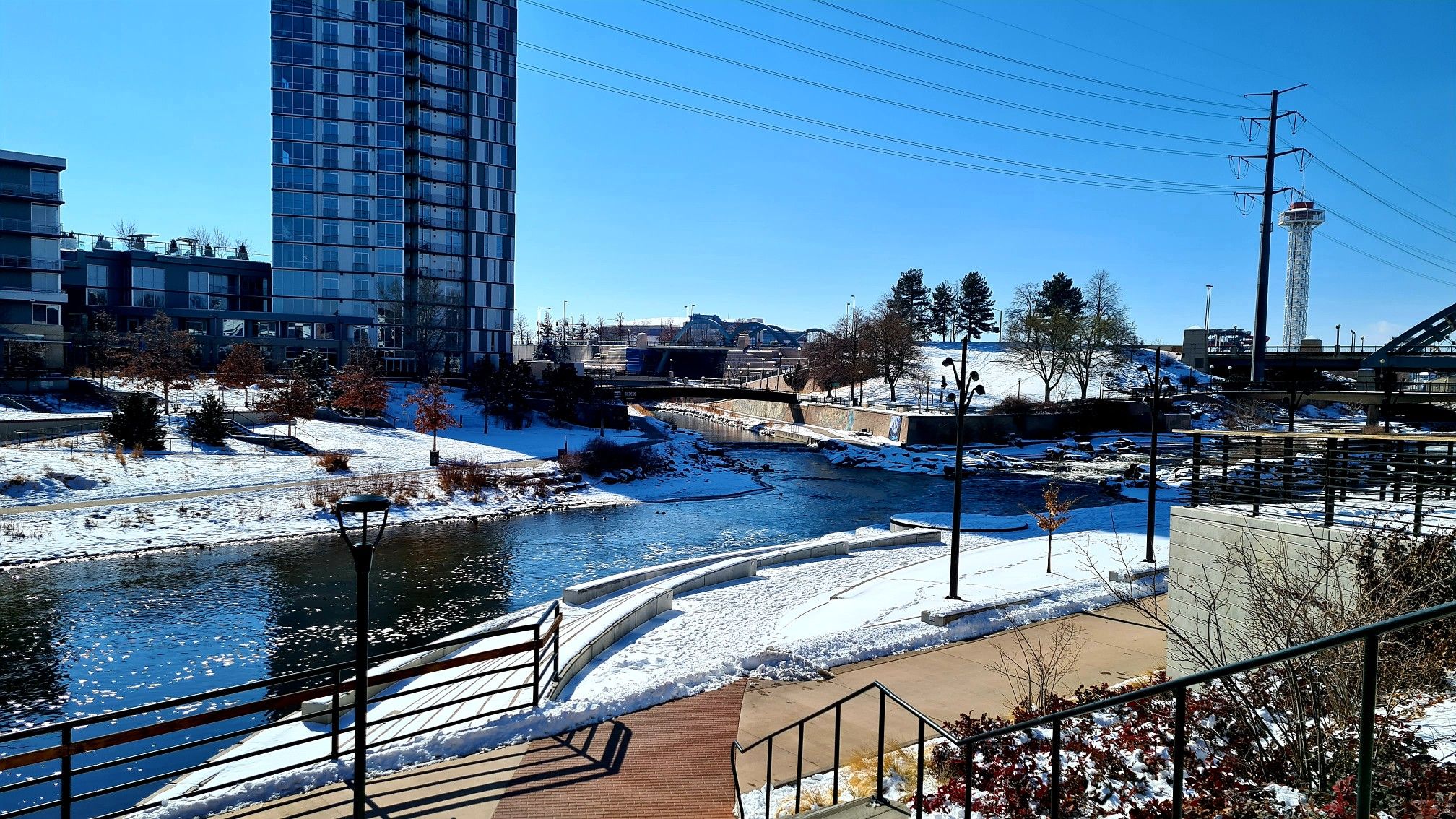 A snowy river flows past tall buildings on a bright, clear winter day. - Confluence Park – Union Station Denver loop from Denver