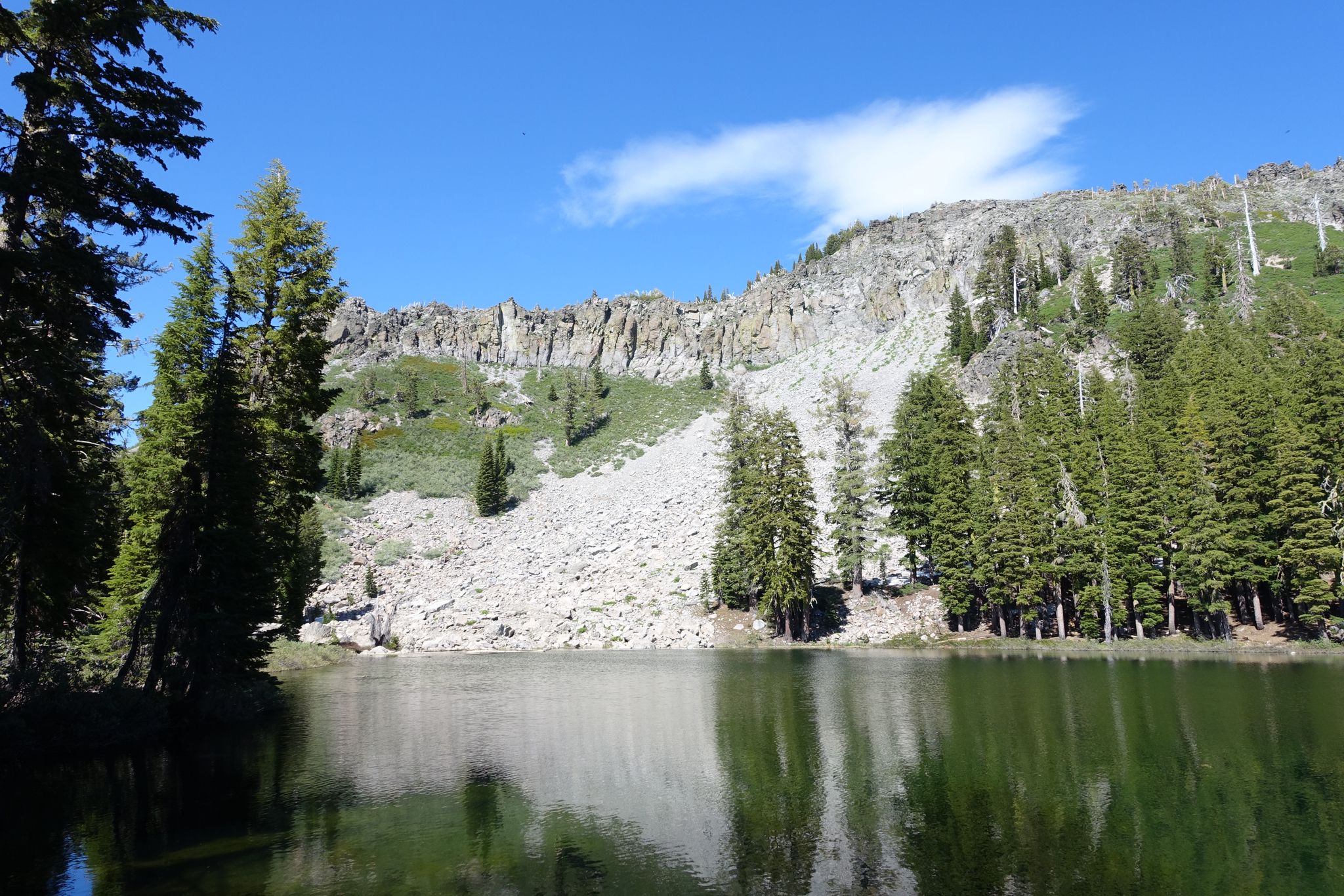 Mountain lake reflecting pine trees and a steep rocky slope under blue sky. - Kaspian Campground to Barker Pass via Barker Pass Road