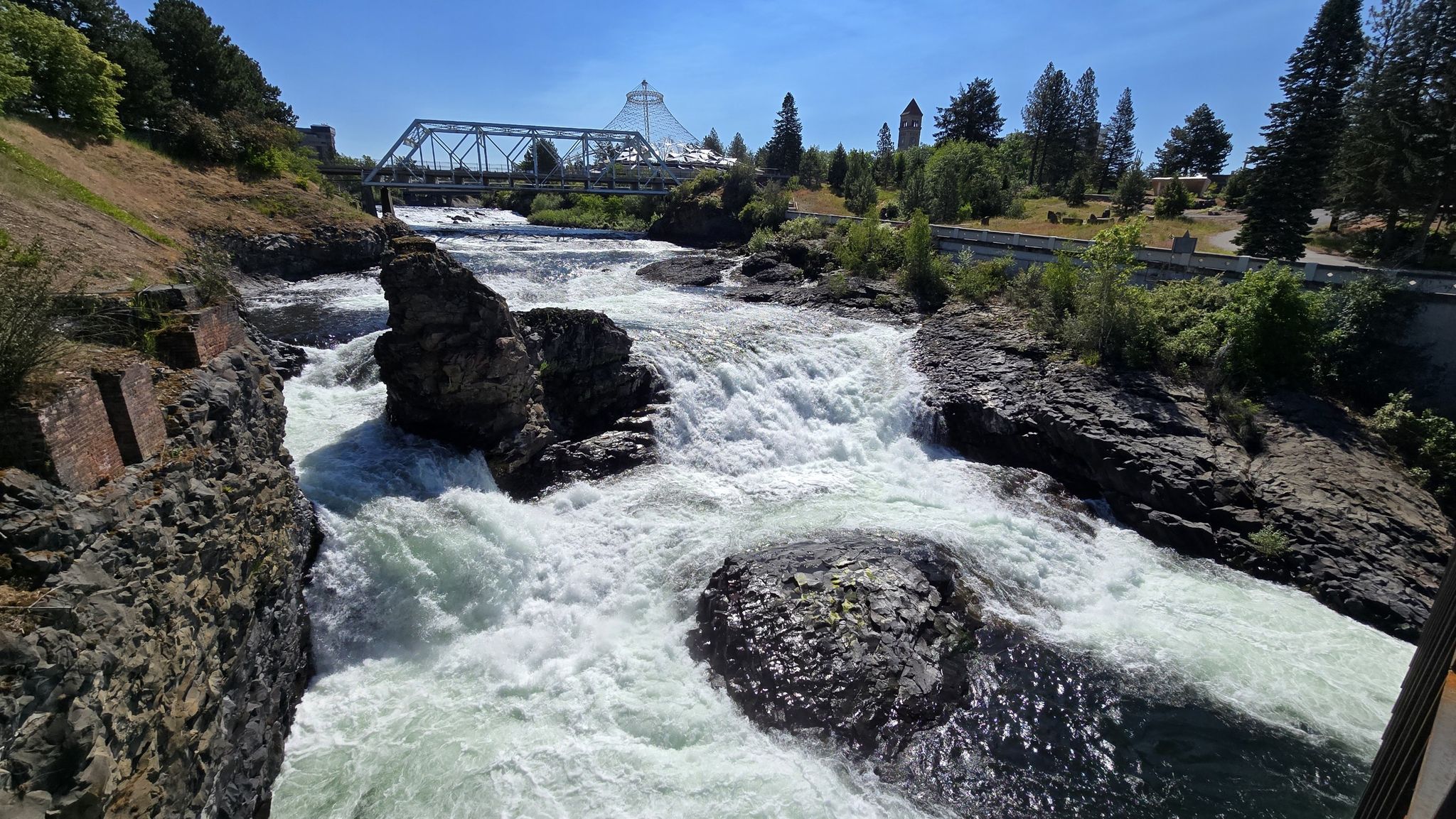 Sunny view of powerful river rapids, a truss bridge, and a large glass structure. - Upper Spokane Falls – Riverfront Park loop from Wyakin Park