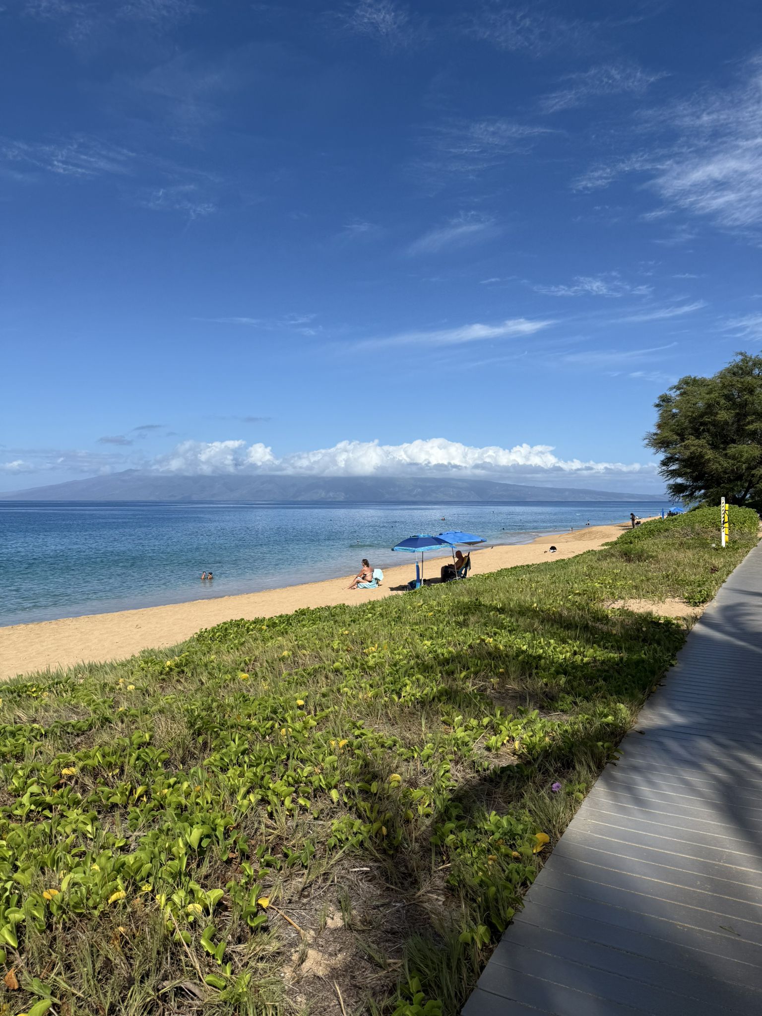 Boardwalk next to a sunny beach with people and distant land. - Kaanapali Beach Path – Kaʻanapali Beach Path loop from Keka'a Beach