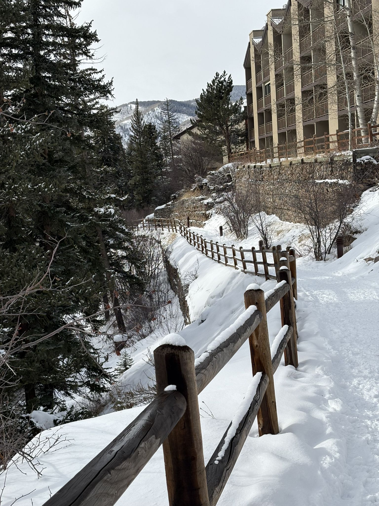 Wooden fence along a snowy path next to evergreen trees and a large building. - Gore Valley Trail