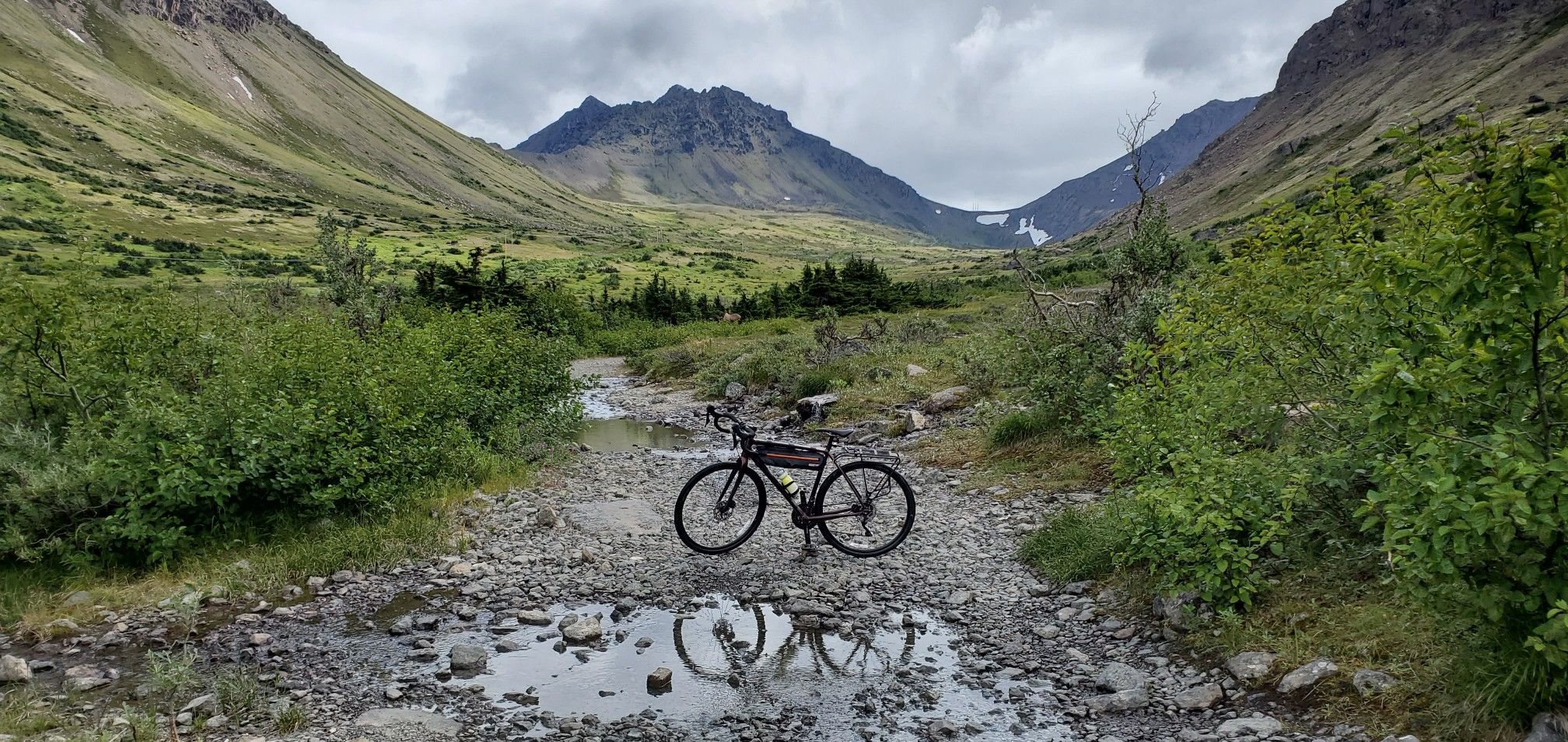 Bicycle on a rocky path with a puddle, framed by green mountains. - Powerline Pass Trail