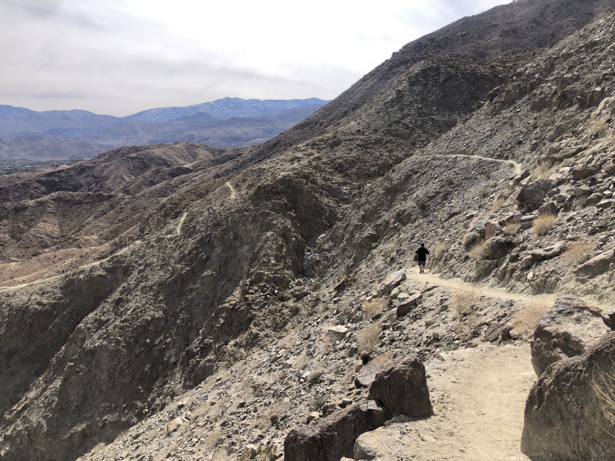 A person hikes on a winding dirt trail along a steep, rocky mountain. - Views of Palm Desert loop from College of the Desert