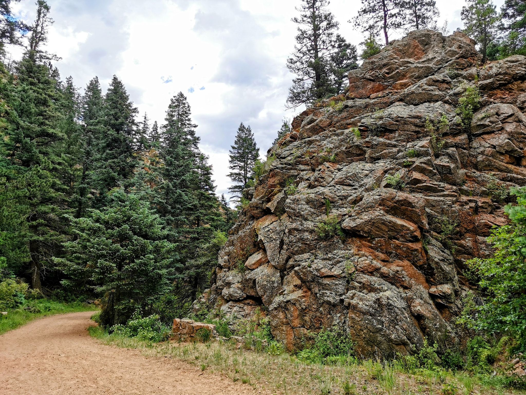 A large, textured rock formation towering over a dirt path and pine trees. - Old Colorado City – Helen Hunt Falls loop from Colorado Springs