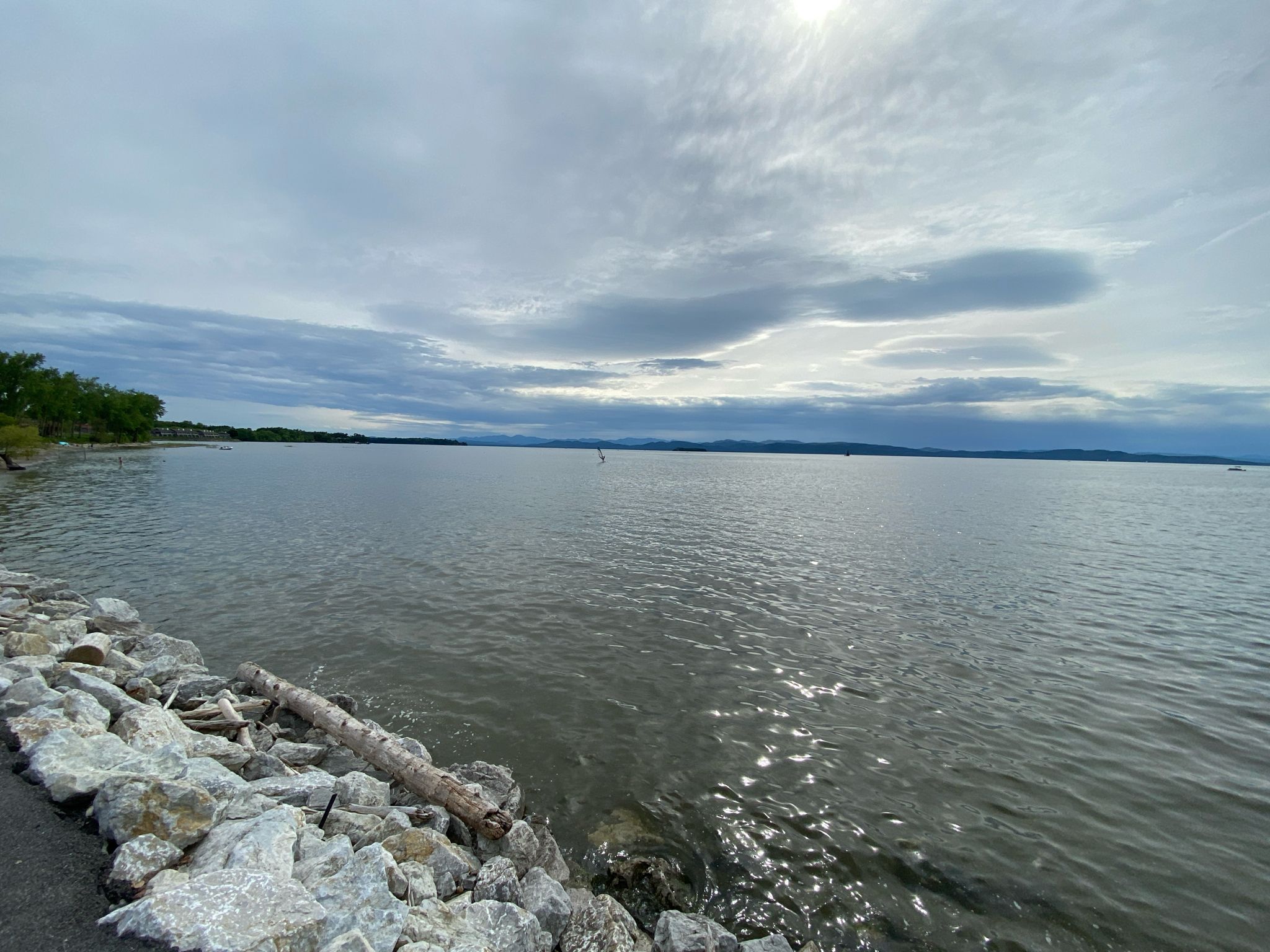 A wide body of water with a rocky shore under a bright, cloudy sky. - Champlain Bikeway