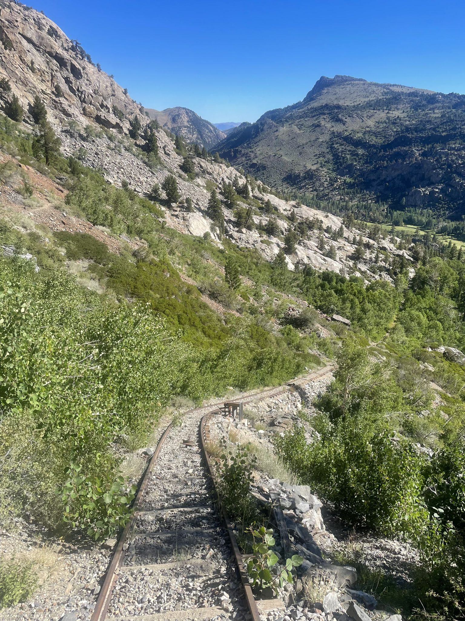 Winding train tracks through a steep, rocky, green mountain valley on a sunny day. - Rush Creek Trail to Agnew Lake