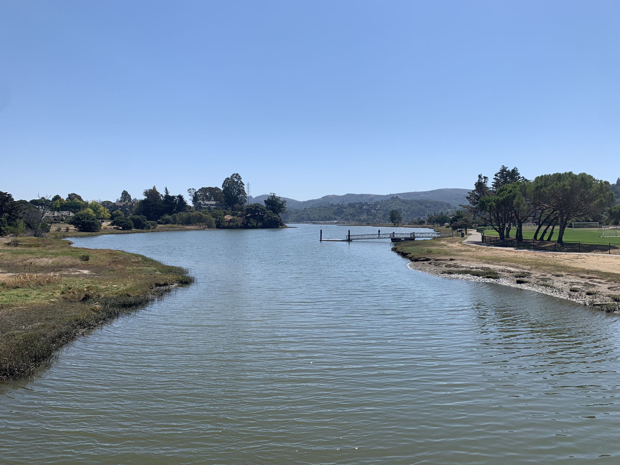 A calm river flows through marshland with distant trees and houses under blue sky. - San Francisco Bay Trail: Paradise Loop