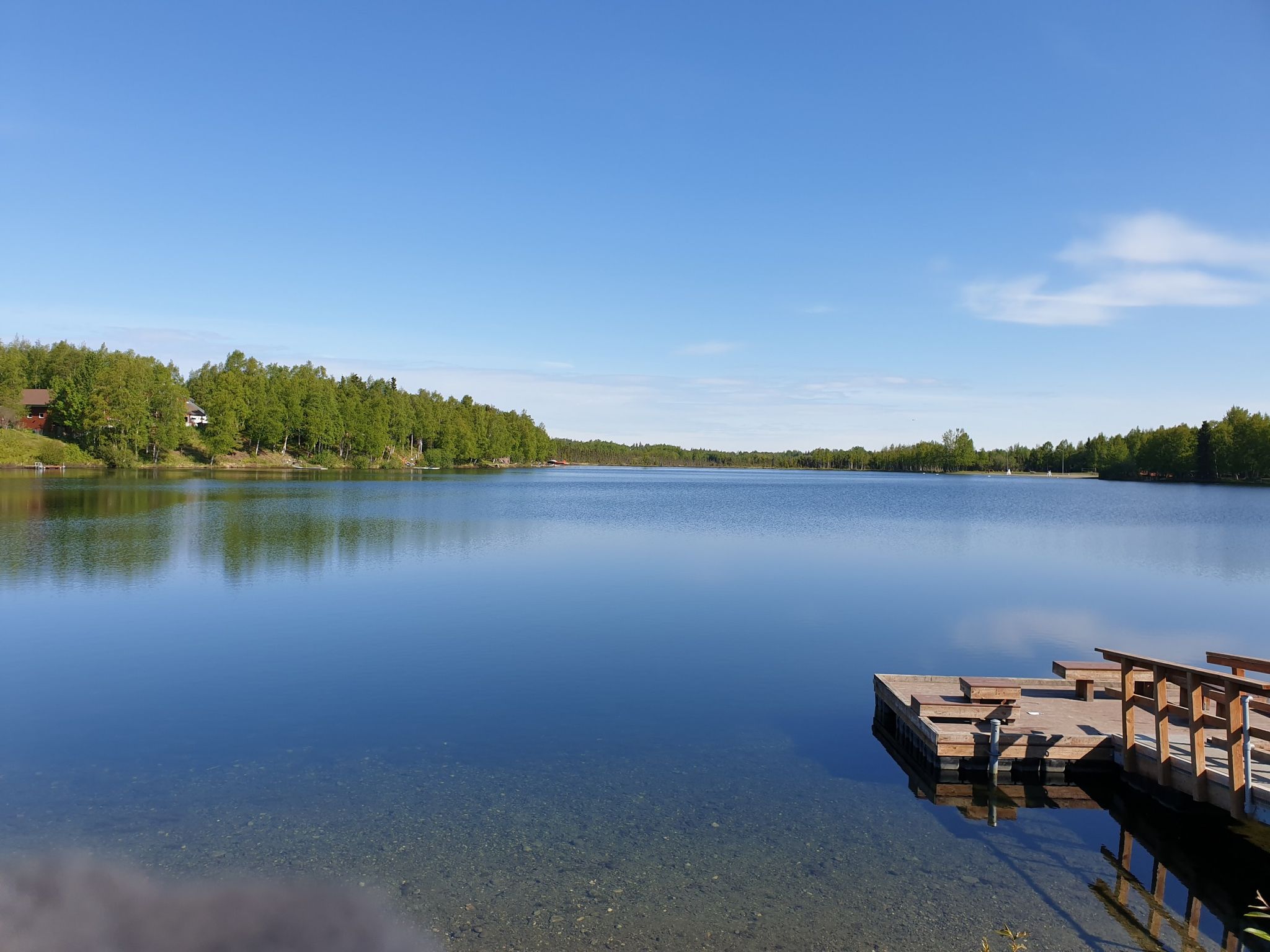 Calm lake with a wooden dock and green trees under a bright blue sky. - Kincaid Chalet – View of Cook Inlet loop from Pollock Homestead