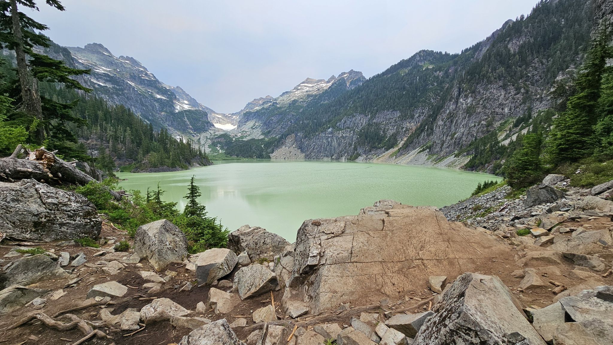 Light green mountain lake surrounded by steep, rocky, forested slopes under an overcast sky. - Blanca Lake Trail
