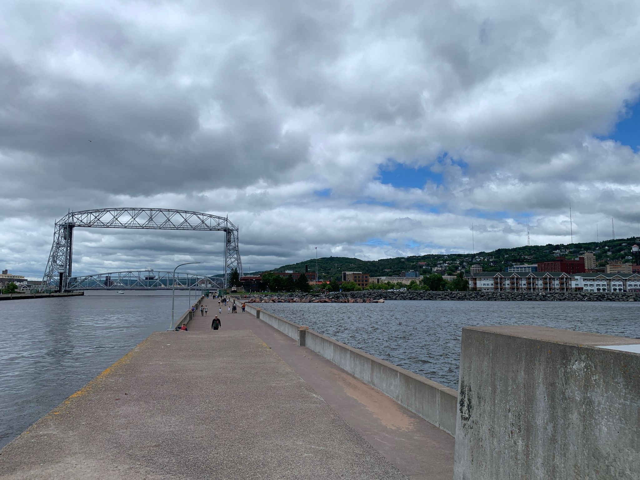 People walk on a pier towards a large steel lift bridge under cloudy skies. - Superior Hiking Trail: Canal Park to Enger Park
