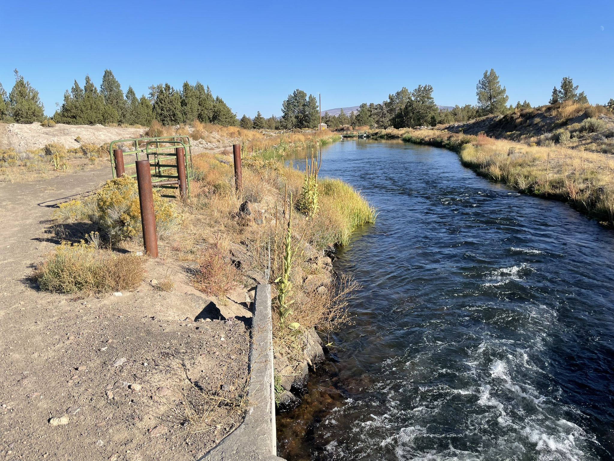 A clear river flows past a dirt path and a wooden fence on a sunny day. - Hiking loop from Tumulus Trailhead