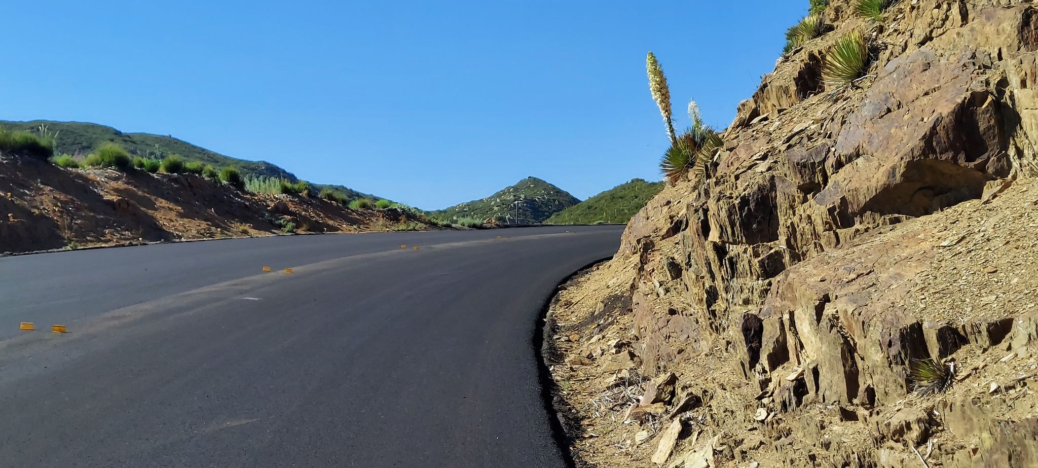 Curved asphalt road next to a steep, rocky hillside under a clear blue sky. - Pine Valley Park – Storm Canyon Overlook loop from Japatul Valley Road Park And Ride