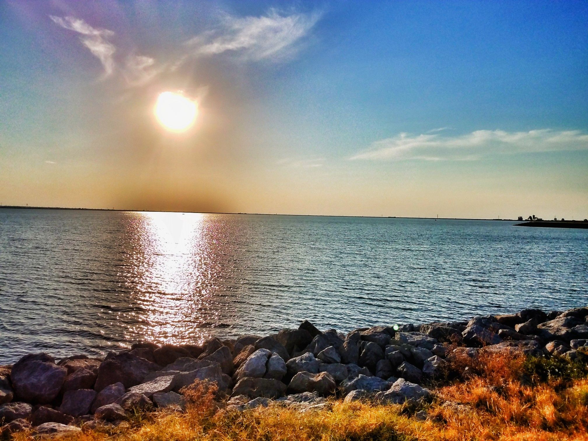 A bright sunset over a calm body of water with a rocky foreground. - Lake Hefner Lighthouse – Bert Cooper Trails loop from Hefner Junior High School