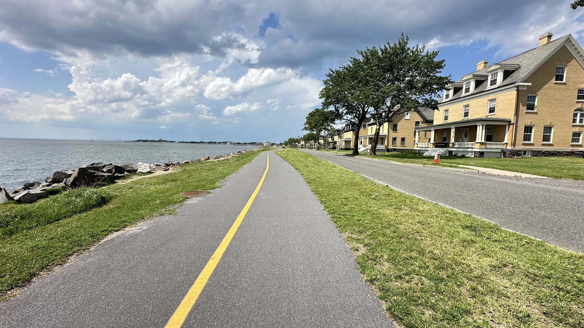 Paved path with yellow line next to water and large buildings under cloudy sky. - Sandy Hook Multi-Use Path