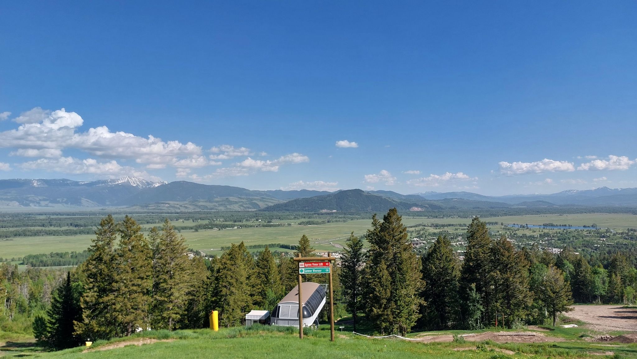 A ski lift structure and evergreen trees overlook a sunny green valley and distant mountains. - Teton Village to Granite Canyon Crossing