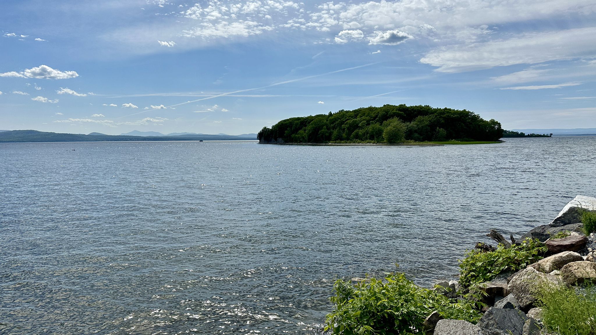 A wide lake with a tree-covered island under a partly cloudy sky. - Island Line Trail