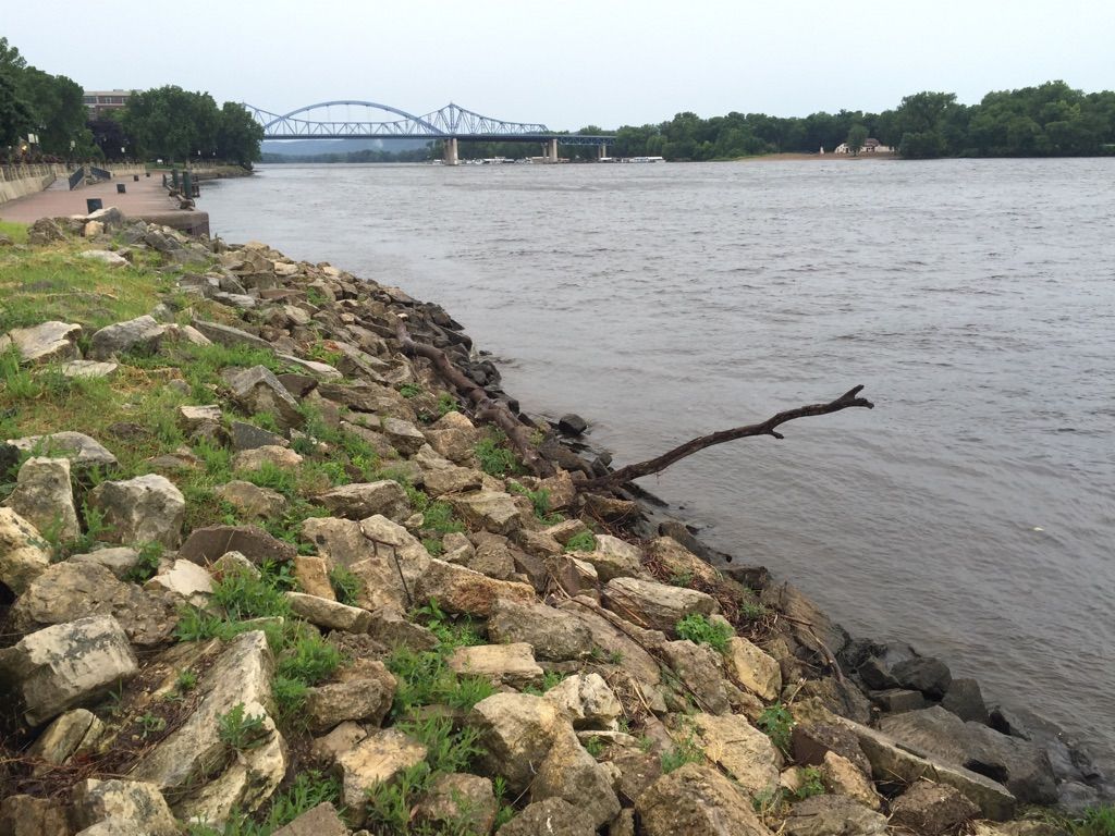 Rocky riverbank, wide river, and distant bridge under an overcast sky. - Riverside Park loop from La Crosse