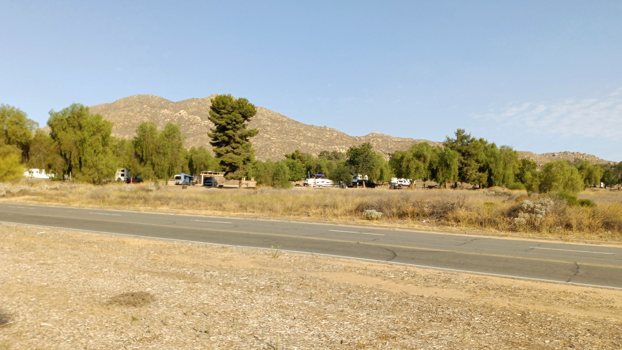 Paved road with trees and RVs, backed by sunny, rocky mountains. - Moreno Beach loop from Lake Perris