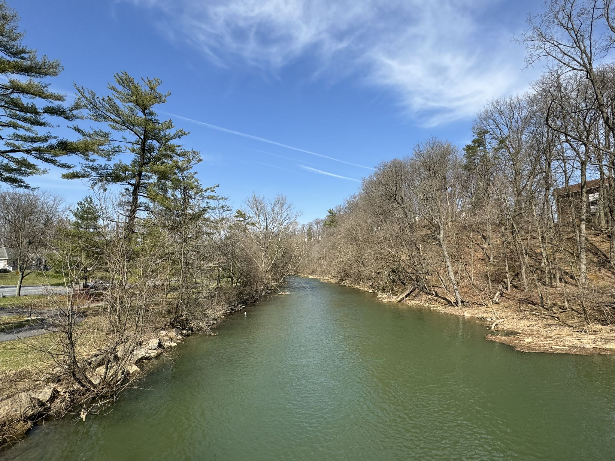 A calm river flows between banks lined with bare trees under a blue sky. - Jordan Creek Greenway and Trexler Park Loop