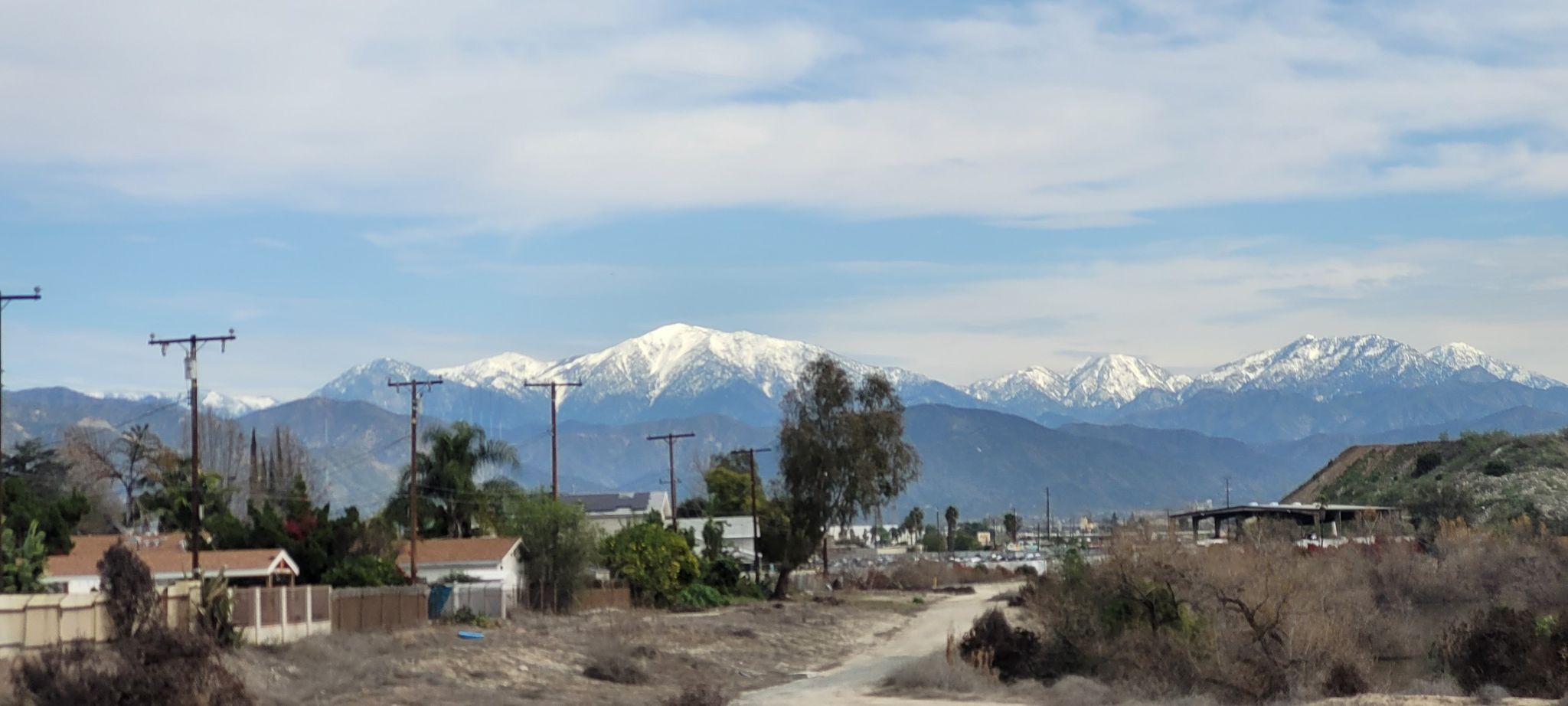 Snow-capped mountains rise behind houses and dry brush under a cloudy sky. - Bosque del Rio Hondo – Rio Hondo Greenway loop from Pio Pico Elementary School