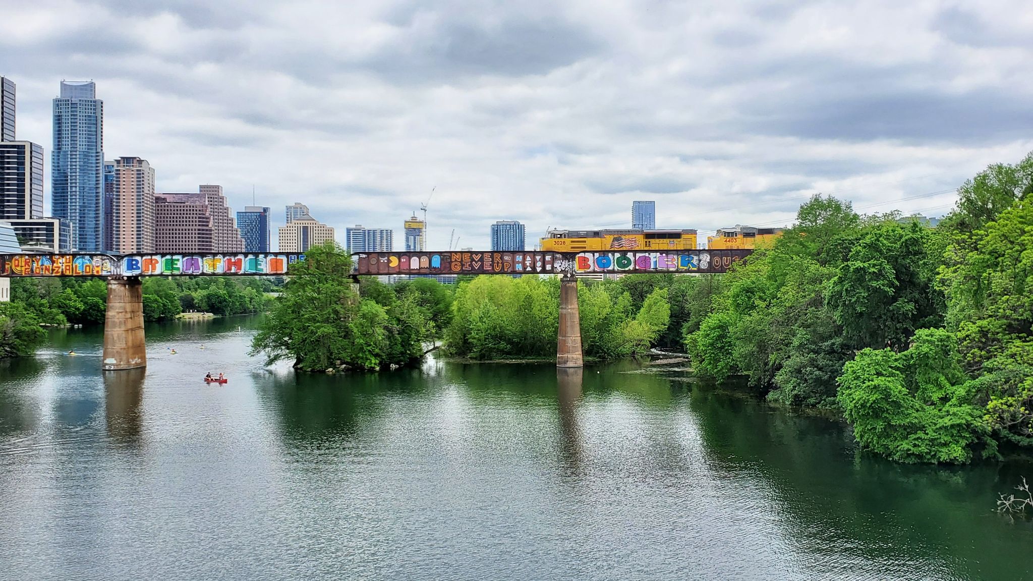 Overcast view of a river, graffiti bridge, city skyline, and kayakers. - Barton Creek Pedestrian Bridge – Lamar Boulevard Bridge loop from Travis