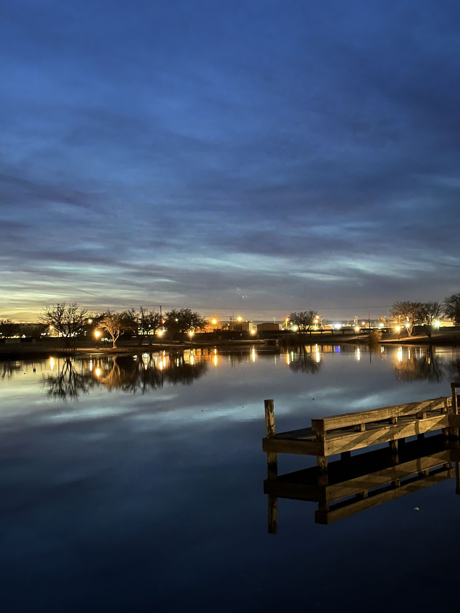 Wooden dock on calm water at dusk, with distant lights reflecting. - Pecos Riverwalk