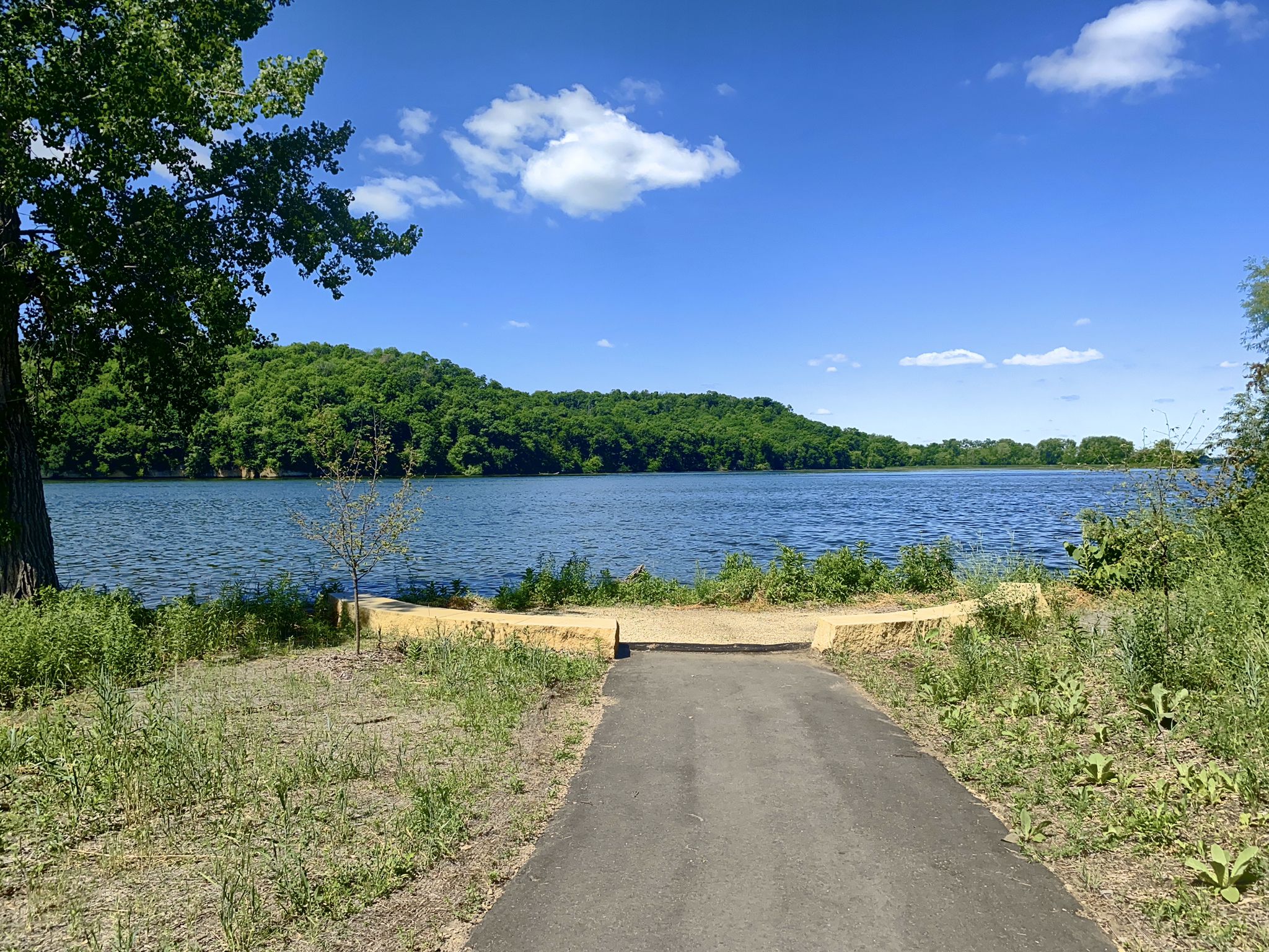 Paved path leading to a lake with forested hills under a blue sky. - Lake Byllesby Dam – Lake Byllesby loop from Cannon Falls