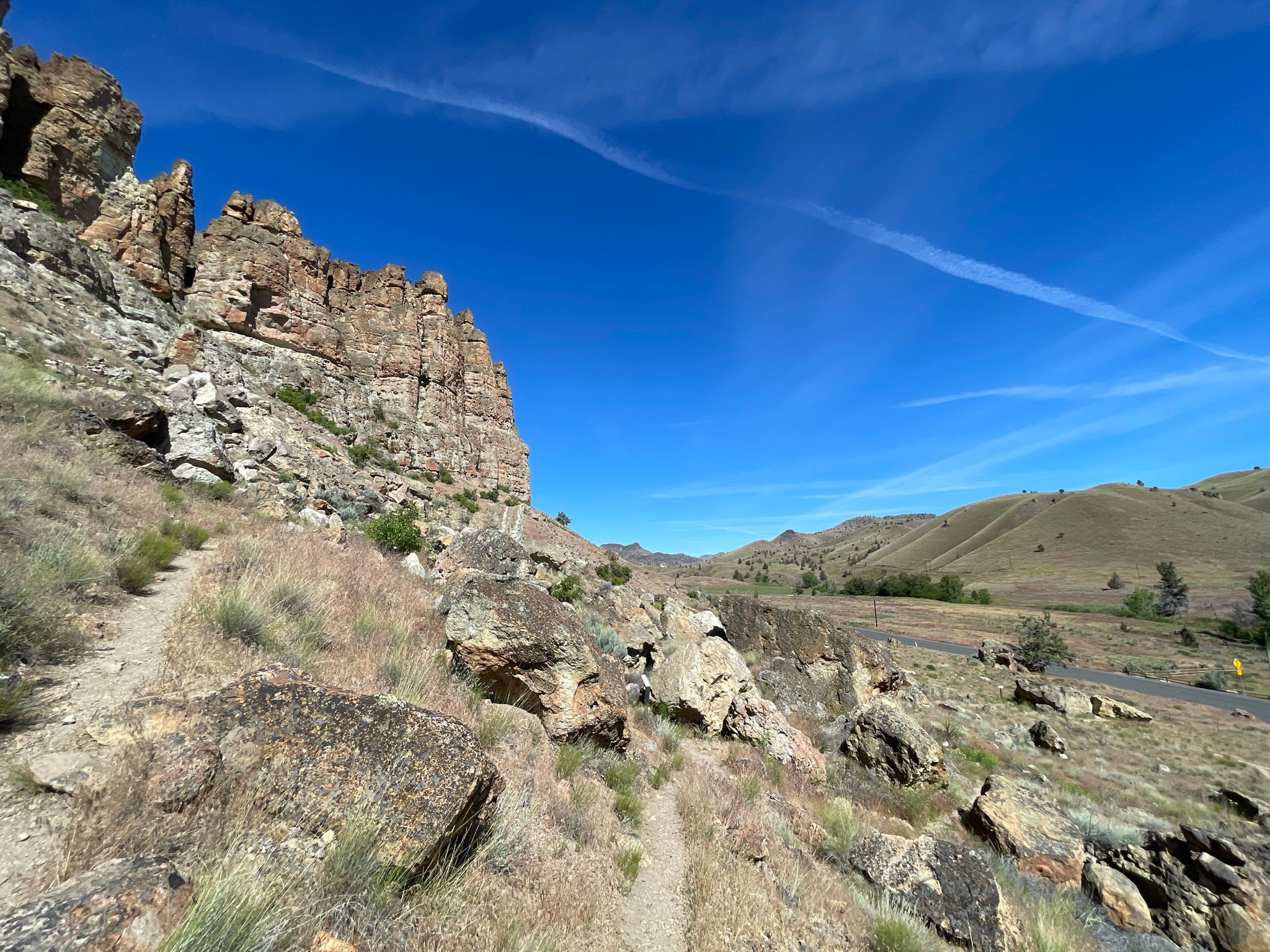 A dirt path winds past rocky cliffs and dry hills under a blue sky. - Geologic Time Trail