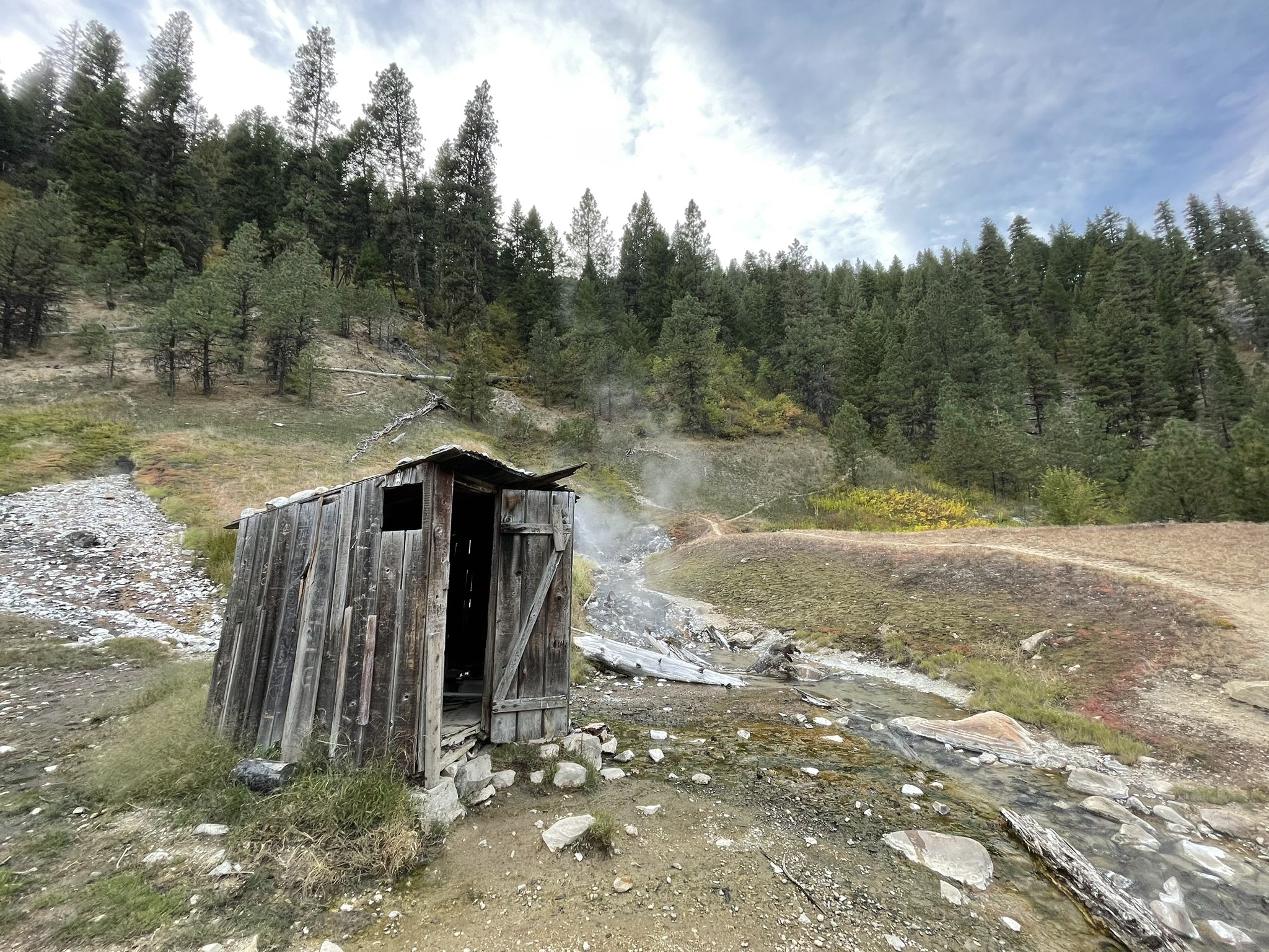 Dilapidated wooden shack sits beside a steaming stream on a grassy, forested hillside. - Bonneville Hot Springs Trail