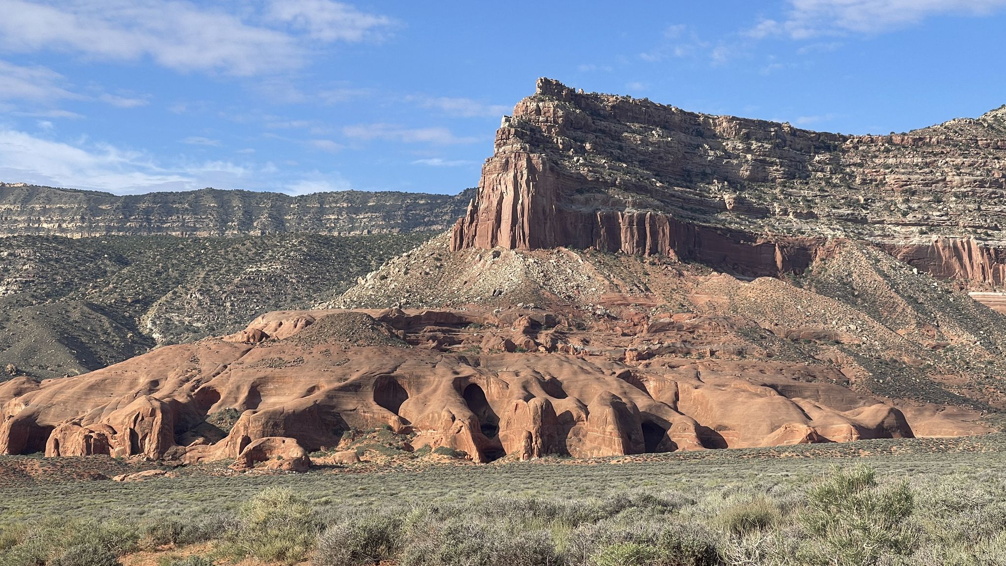 Sunny desert landscape with a massive red rock cliff and eroded base. - Hole-in-the-Rock OHV Road