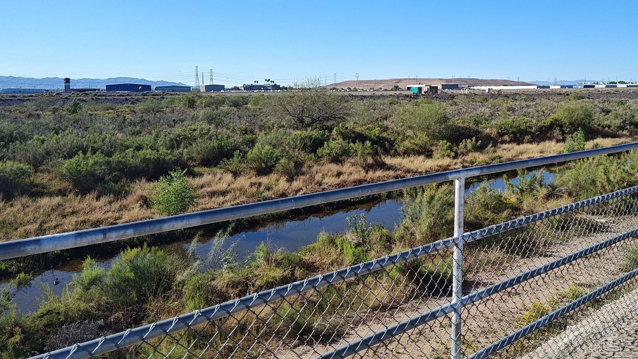 A chain-link fence overlooks a small stream through desert vegetation with distant buildings. - State Farm Stadium - Home of the Cardinals loop from Youngtown