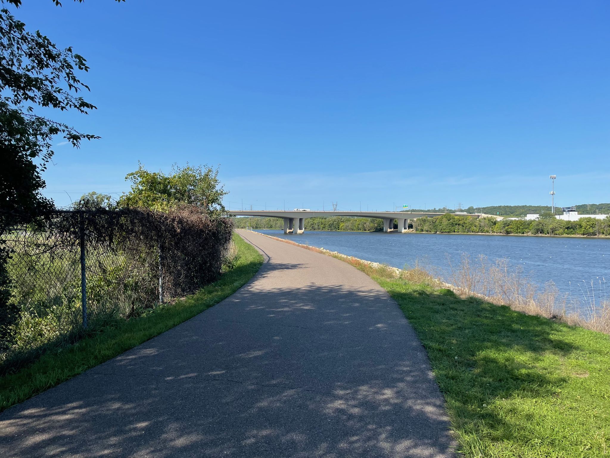 Paved path curves alongside a river with a large bridge in the distance. - Wakota Trailhead loop from Saint Paul Park