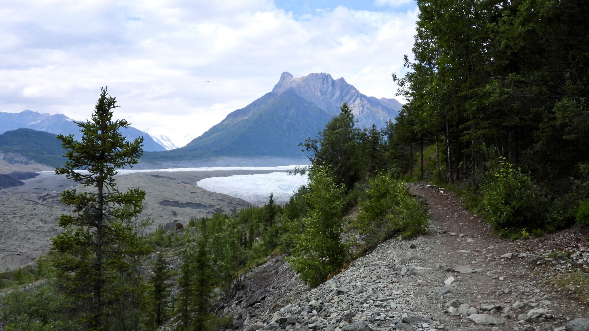 A dirt path overlooks a valley with a lake and mountains under a cloudy sky. - West Kennecott Glacier Trail