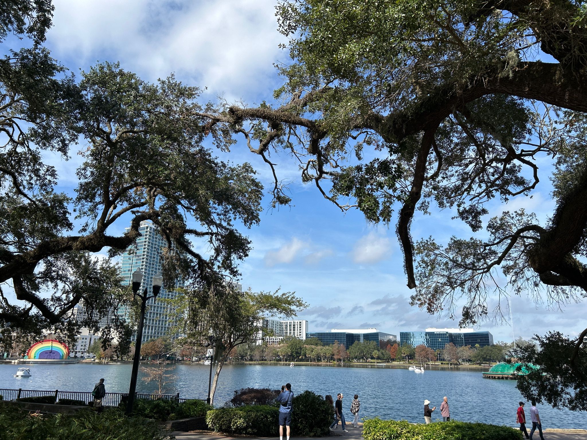 People gather by a lake framed by trees, with a city skyline. - Lake Eola Fountain – Baldwin park and bike trail loop from Church Street