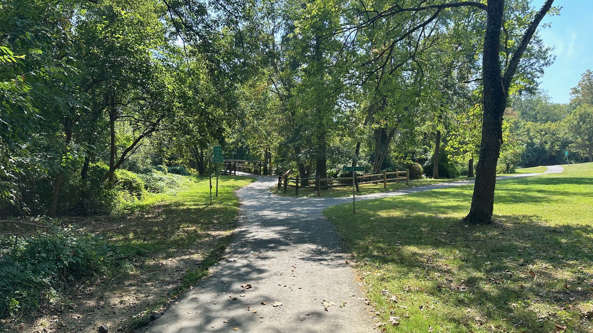 A paved path winds through a sunny park with green grass and trees. - Meadowbrook Stables loop from Forest Glen