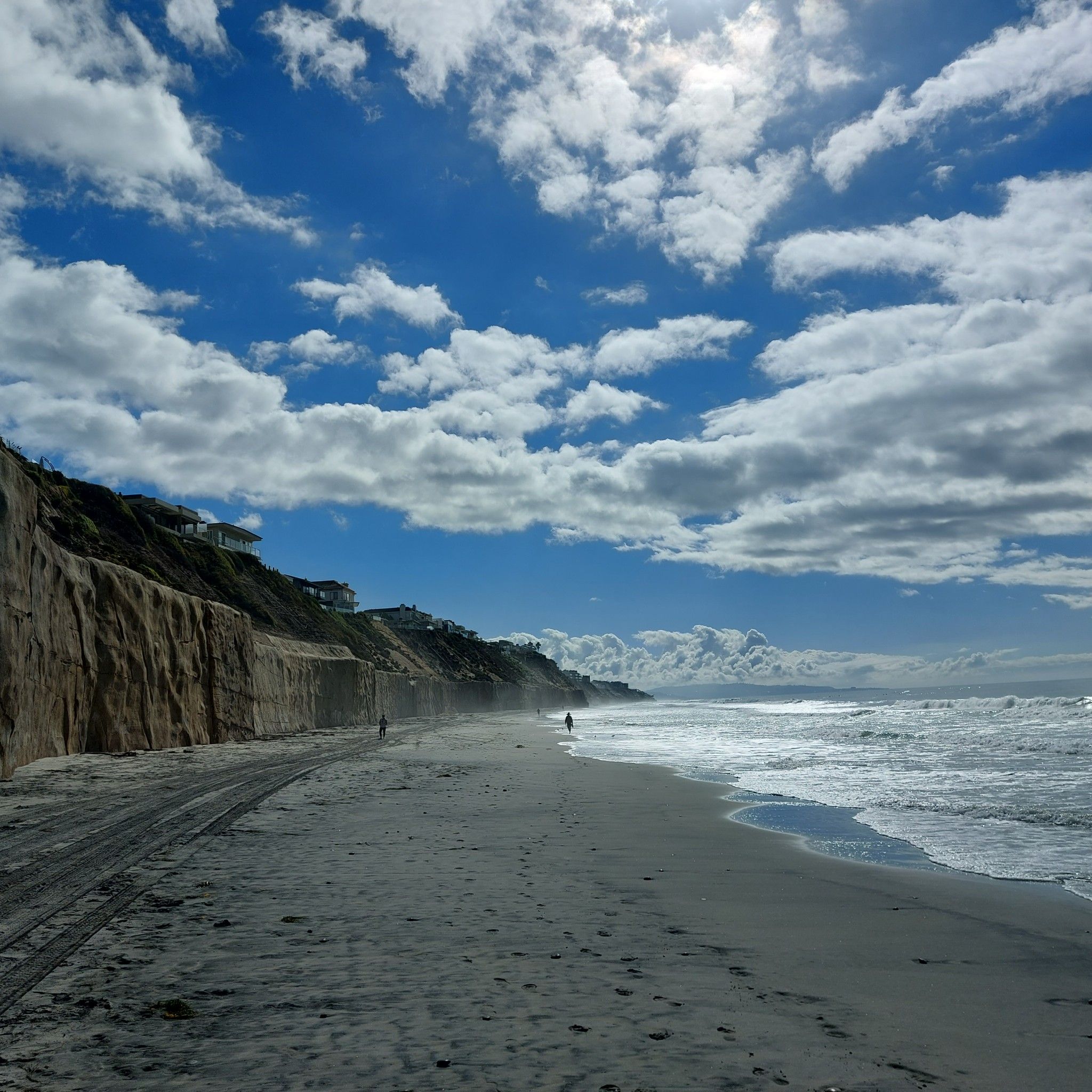Sunny beach with ocean waves, tall cliffs, and distant walkers under cloudy sky. - Coastal Rail Trail