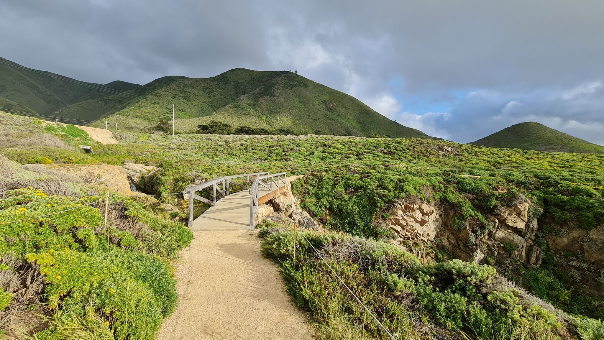 Dirt path leads to a wooden bridge, green hills under a cloudy sky. - Garrapata Bluff Trail