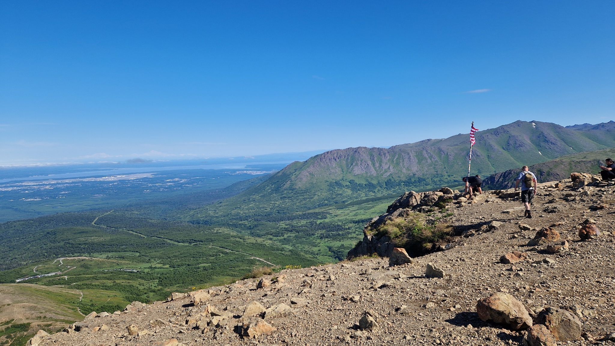 Hikers on a rocky mountain ridge overlooking a vast green valley and distant water. - Flattop Sunnyside Trail