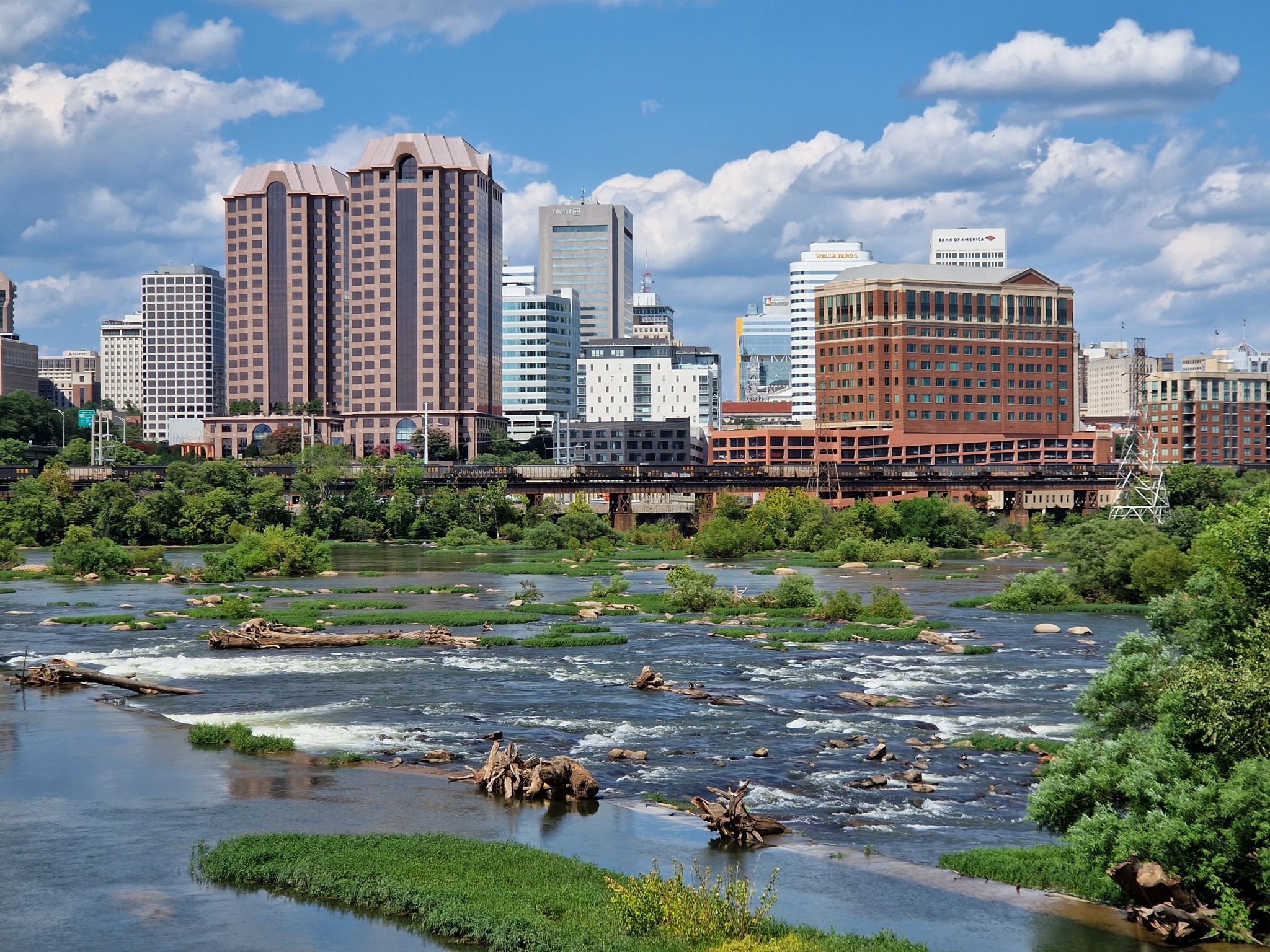 River with rapids and green islands, backed by a distant city skyline. - Manchester Floodwall and Slave Trail