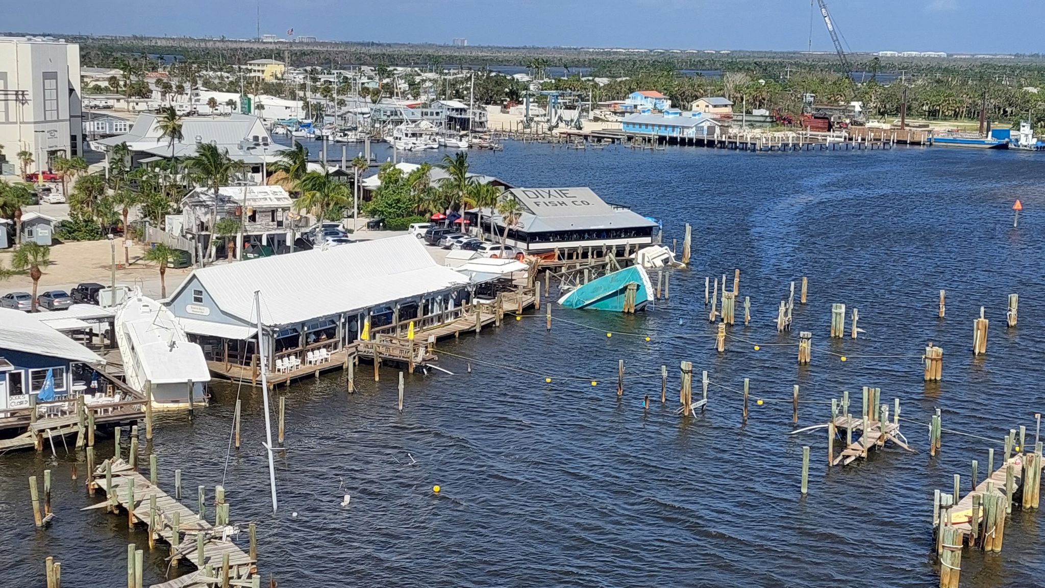 Aerial view of waterfront buildings, damaged docks, and sunken boats under bright sun. - Times Square, Fort Myers Beach – View of Estero Boulevard loop from Smugglers Cove - adventure golf