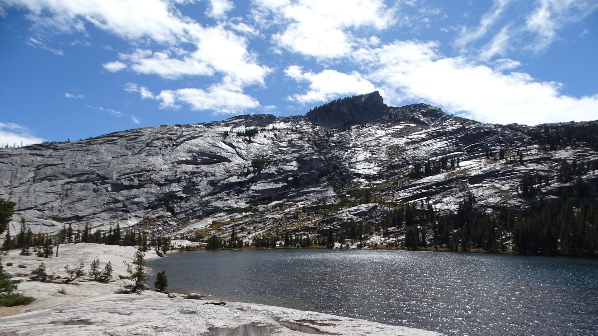 A dark lake at the base of a massive, rocky mountain under cloudy skies. - Cathedral Lakes Trail