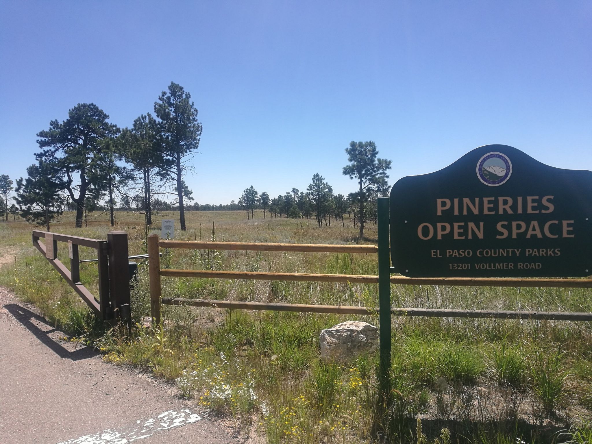 A green sign and wooden fence stand beside a sunny open field. - The Pineries Open Space loop