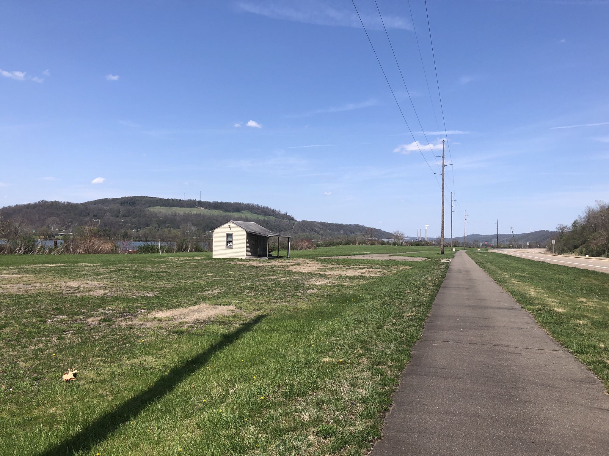 A small shed sits in a sunny green field beside a paved path. - Wheeling Heritage Trail