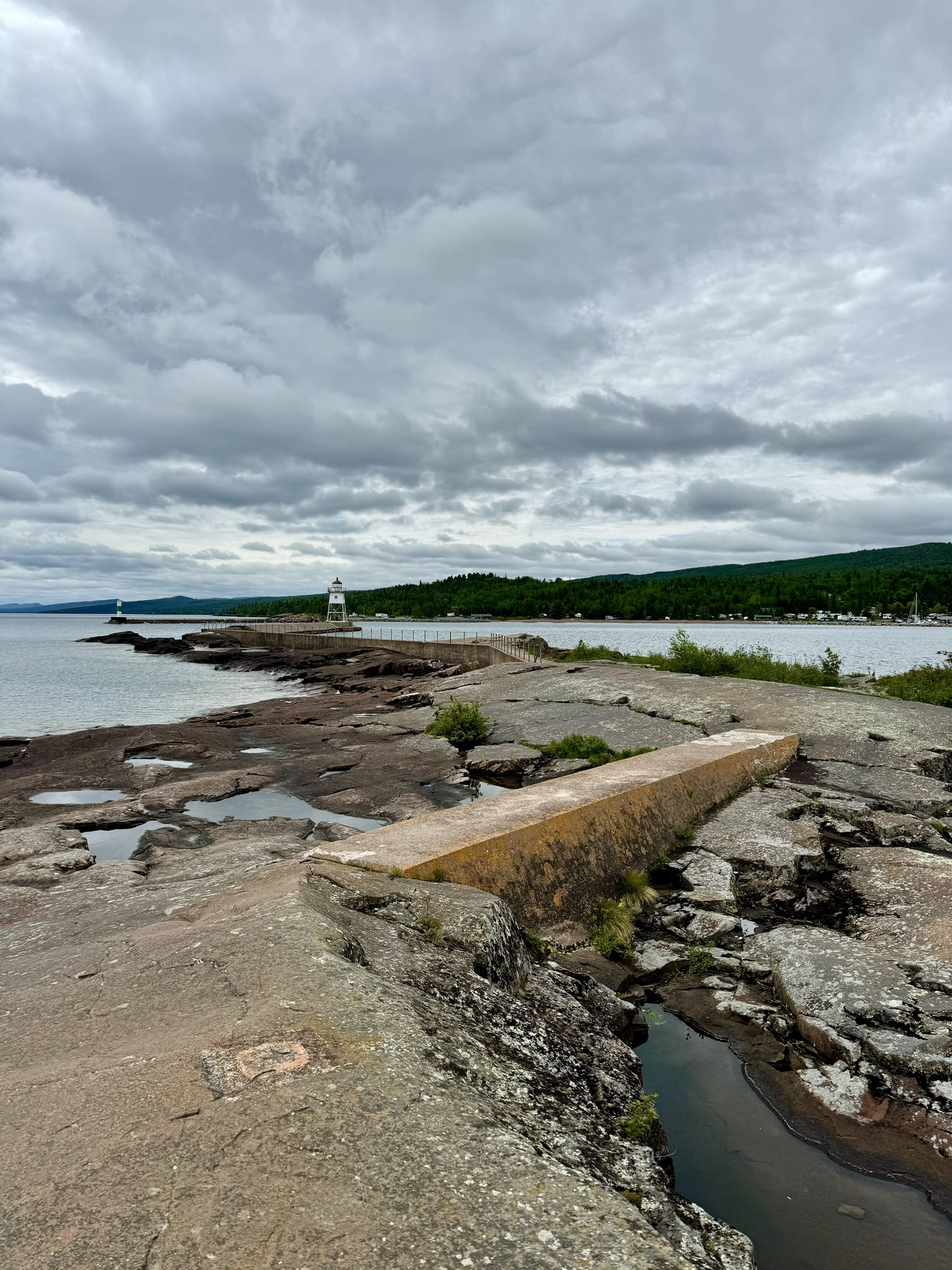 Rocky shoreline and concrete path under an overcast sky, distant lighthouse. - Artists' Point Trail
