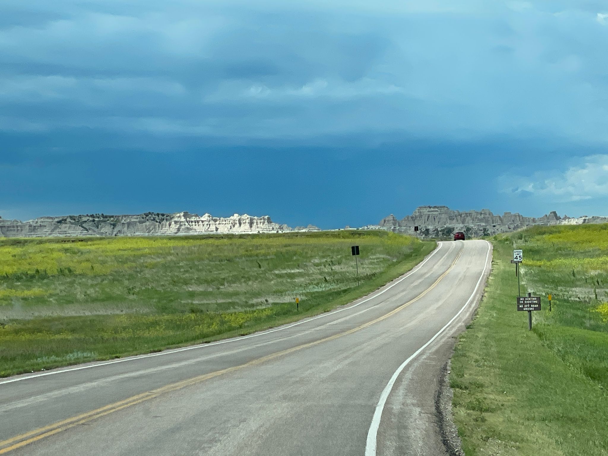 Road winding through green fields towards distant badlands under a cloudy sky. - Northeast Loop