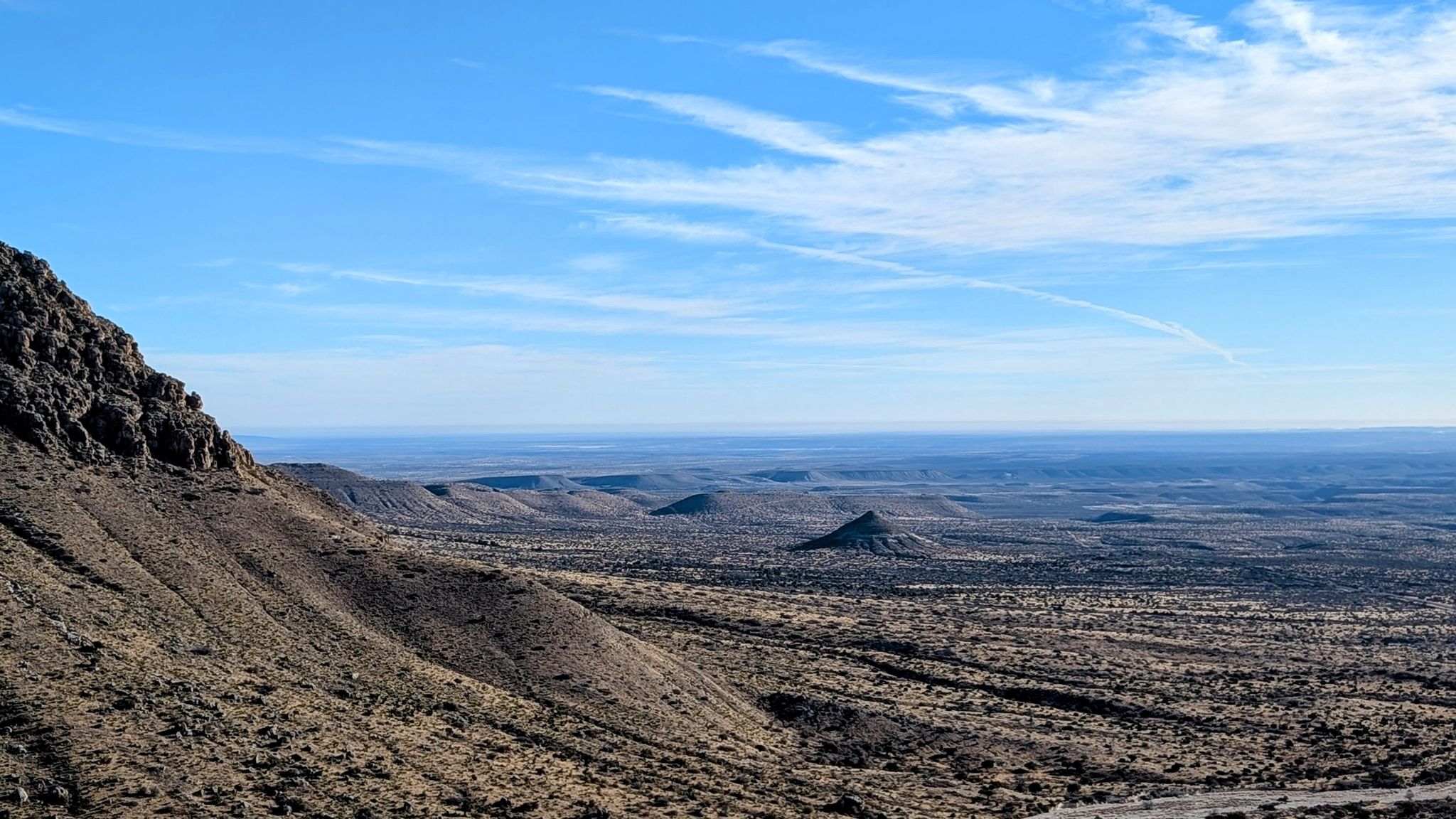 Vast desert landscape with a rocky mountain and distant hills under a blue sky. - Guadalupe Peak Trail