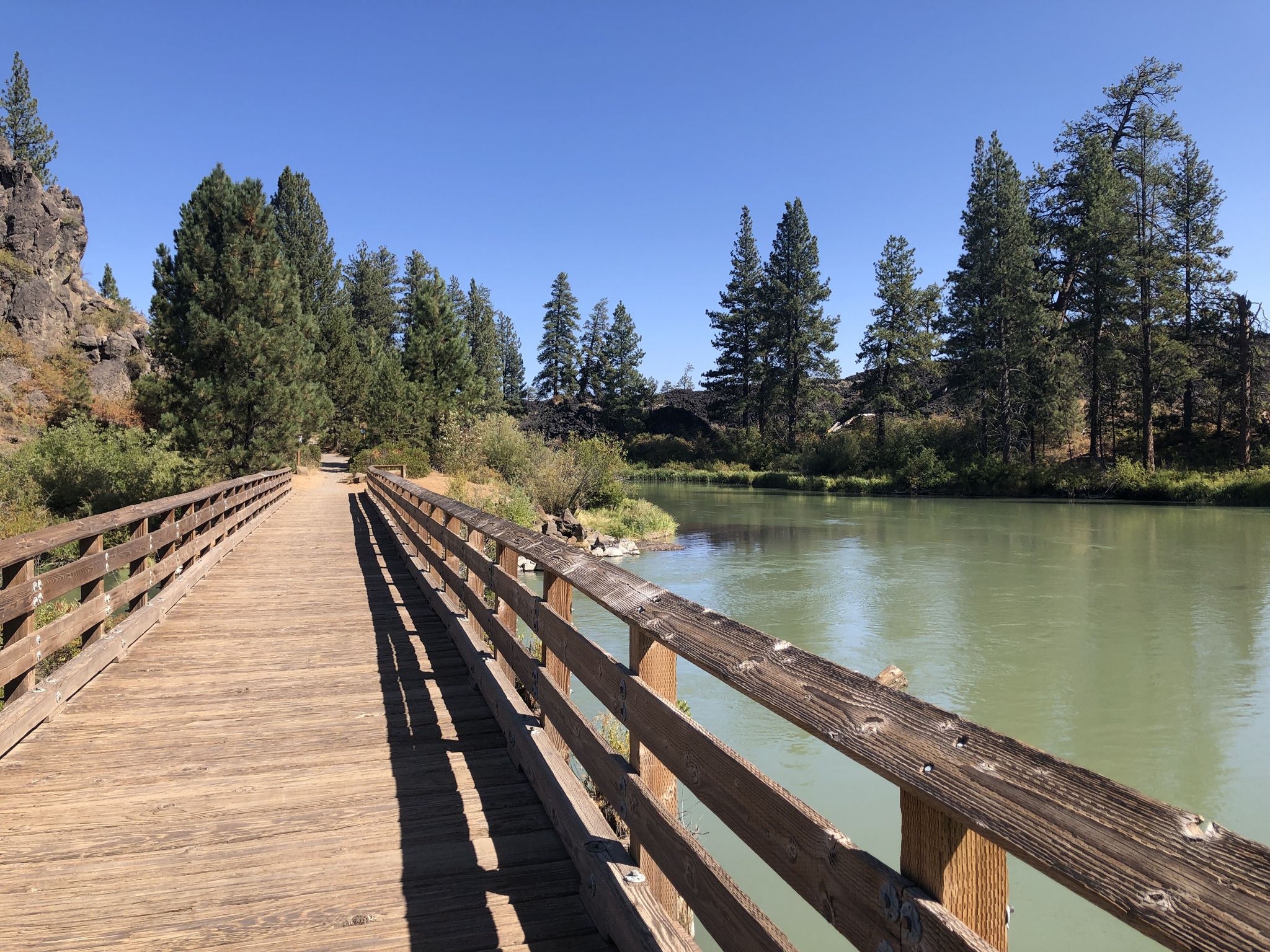 Wooden boardwalk next to a calm, green-tinted river under clear sky. - Trail of the Molten Land – Deschutes River Bridge loop from Lava Butte