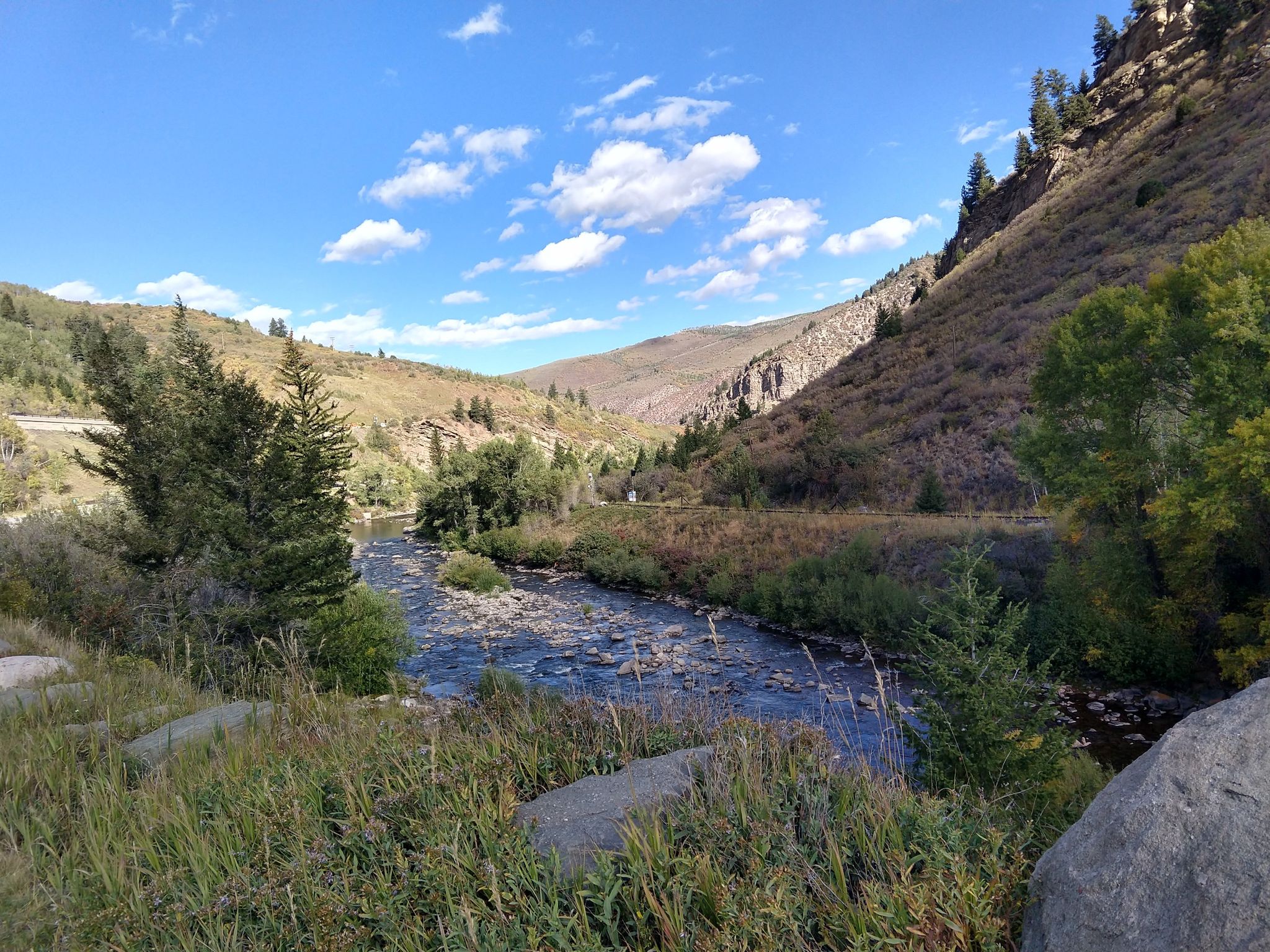 A river flows through a sunny mountain valley with green trees and blue sky. - Gore Valley Trail