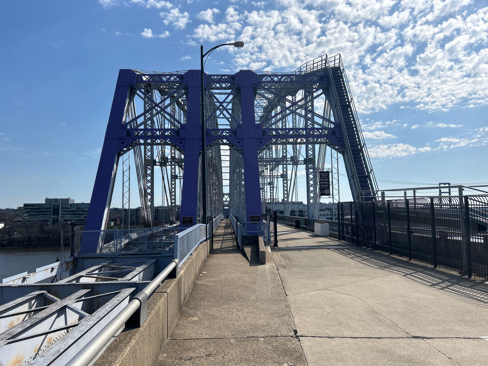 A blue metal bridge with an empty pedestrian walkway under a cloudy sky. - Ohio River Trail – Cincinnati Flying Pig Statues loop from Salem Heights