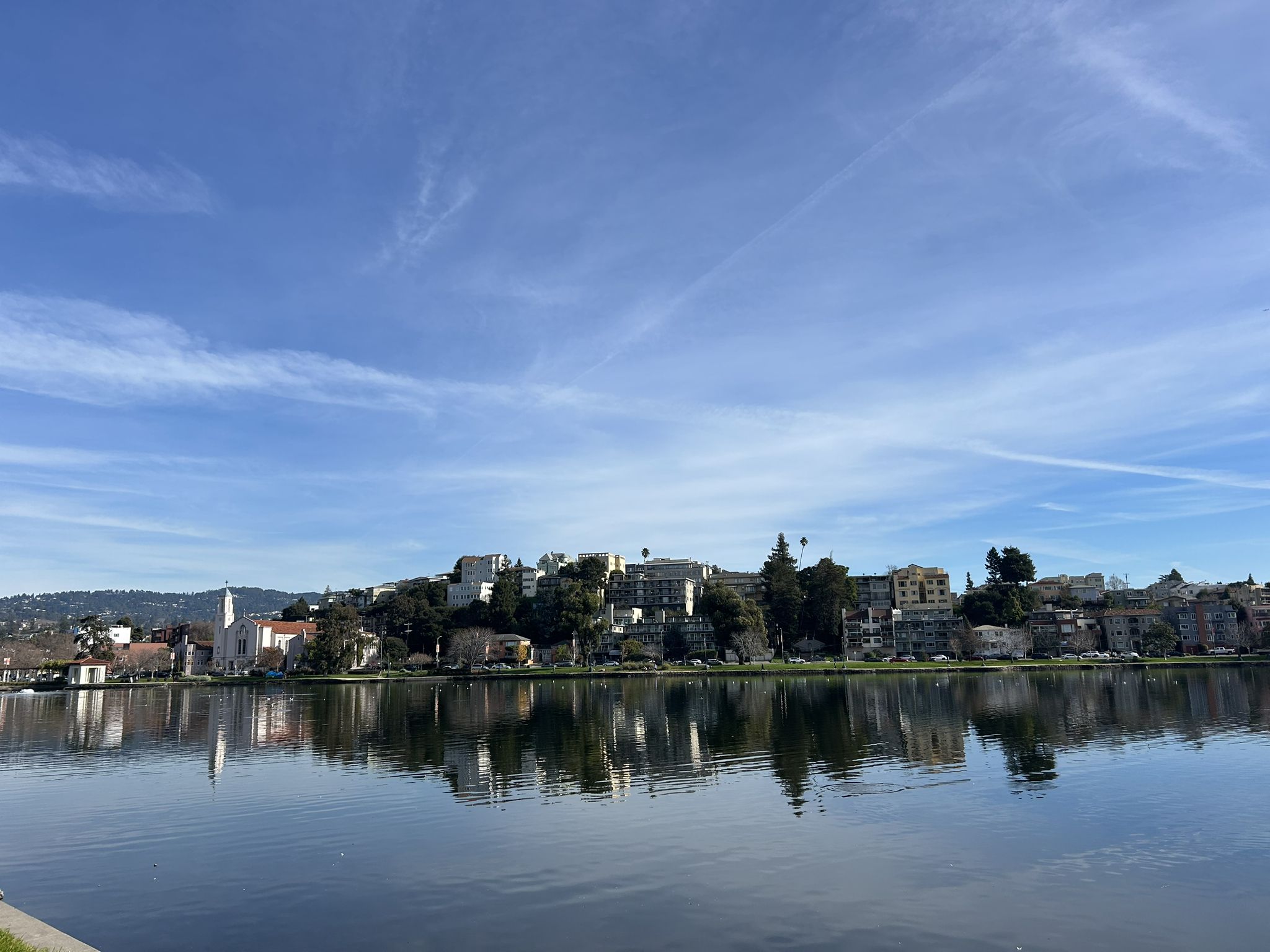 A city skyline reflected in calm water under a bright blue sky with wispy clouds. - Lakeshore Park loop around Lake Merritt